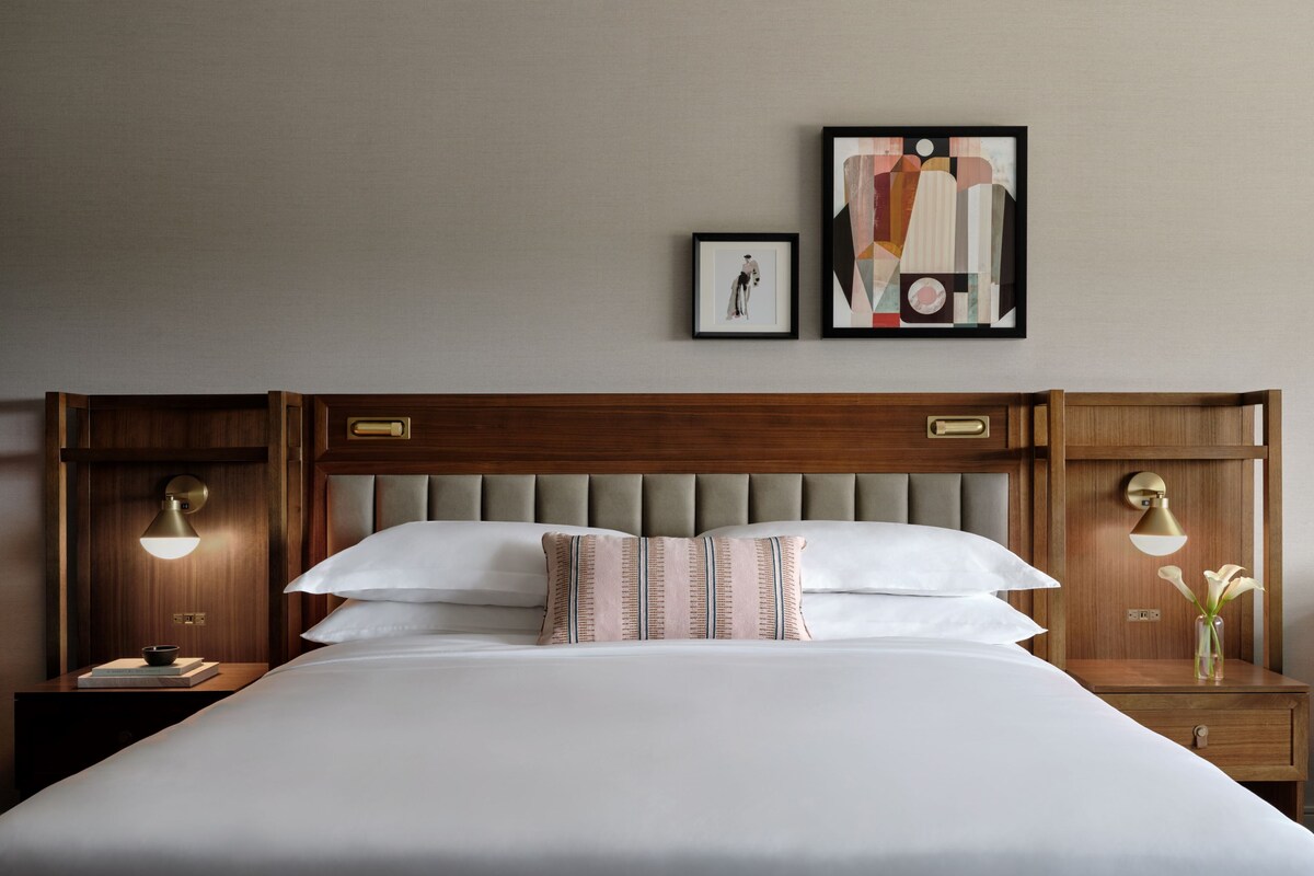 Una cama bien hecha con cabecera de madera, dos mesitas de noche con lámparas y dos cuadros en la pared.