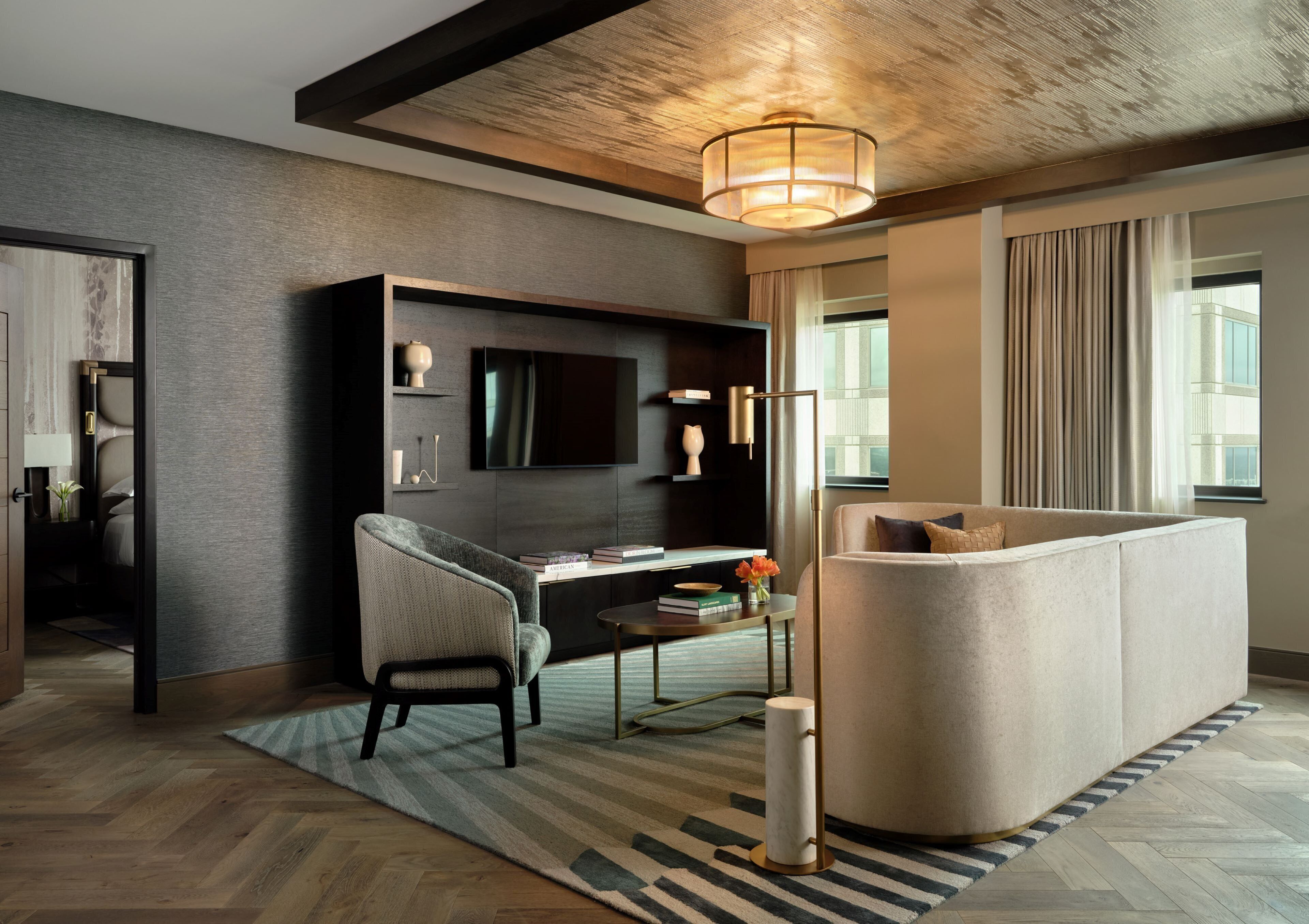 A modern hotel room with a dark wood floor, a large sofa, a desk with a chair, a flat-screen TV, and a large window with curtains.