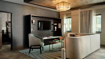 A modern hotel room with a dark wood floor, a large sofa, a desk with a chair, a flat-screen TV, and a large window with curtains.