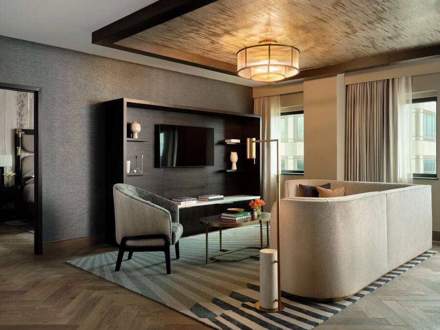 A modern hotel room with a dark wood floor, a large sofa, a desk with a chair, a flat-screen TV, and a large window with curtains.