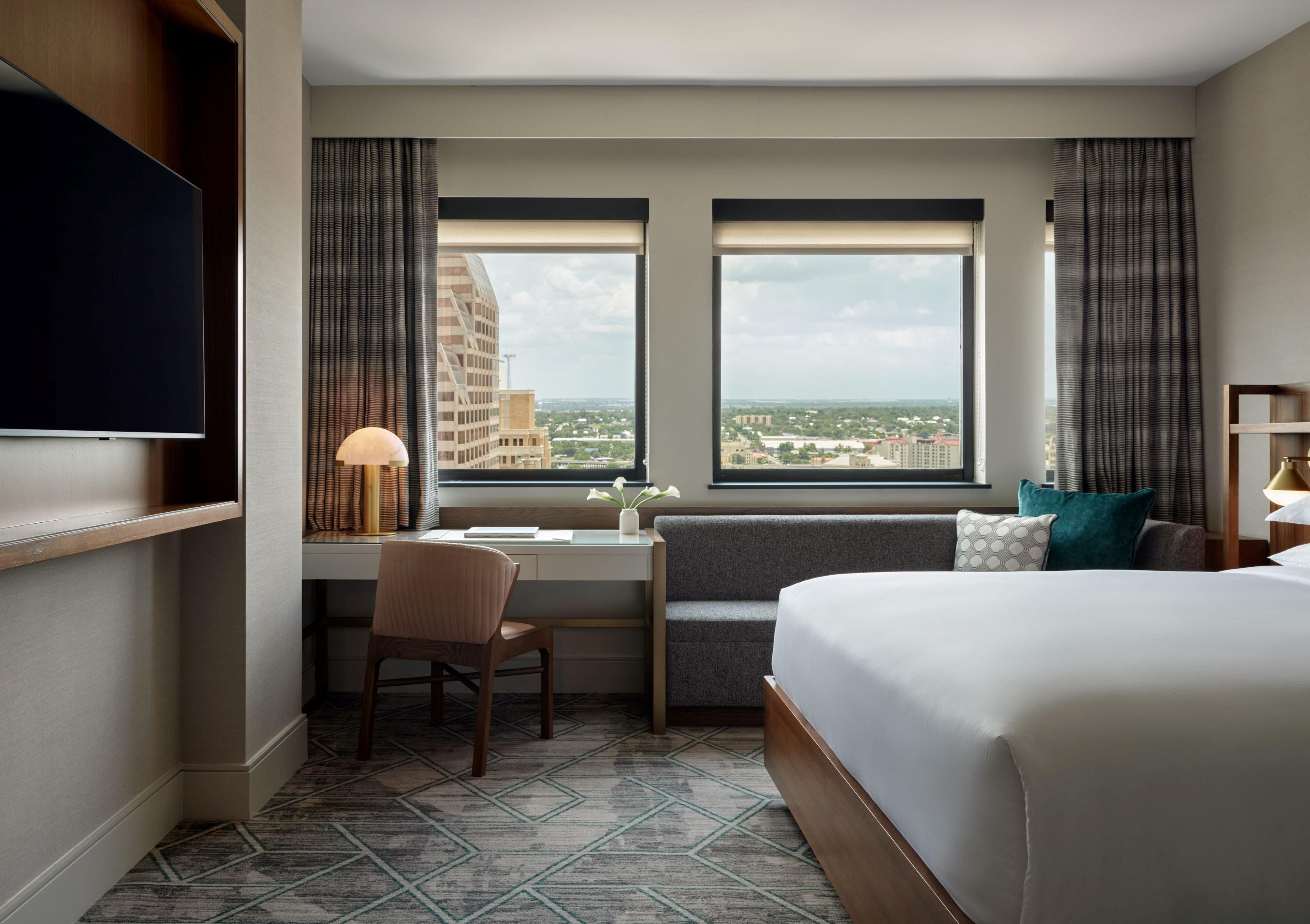 A hotel room with a bed, desk, chair, two windows with curtains, a TV, and a view of buildings.