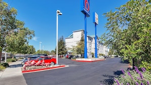 A parking lot with a blue sign, a white building, and a red car. - Motel 6 Belmont, CA - San Francisco - Redwood City (Belmont)
