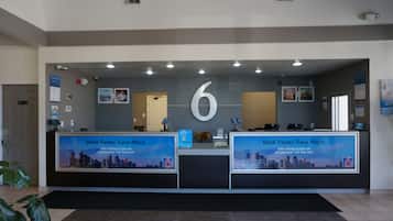 A hotel reception desk with a large number '6' above it, two computer monitors, a reception desk, and a cityscape view.