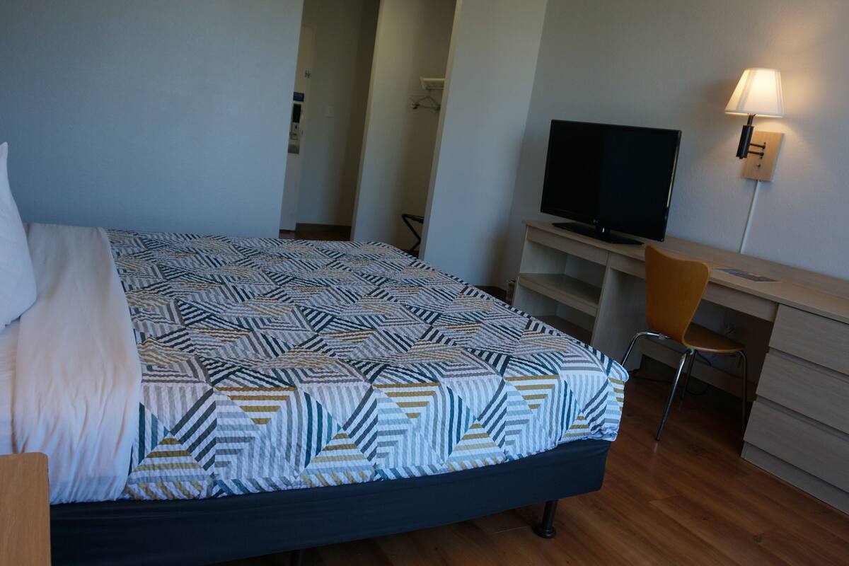 A hotel room with a bed, a desk with a TV, a chair, and a lamp.