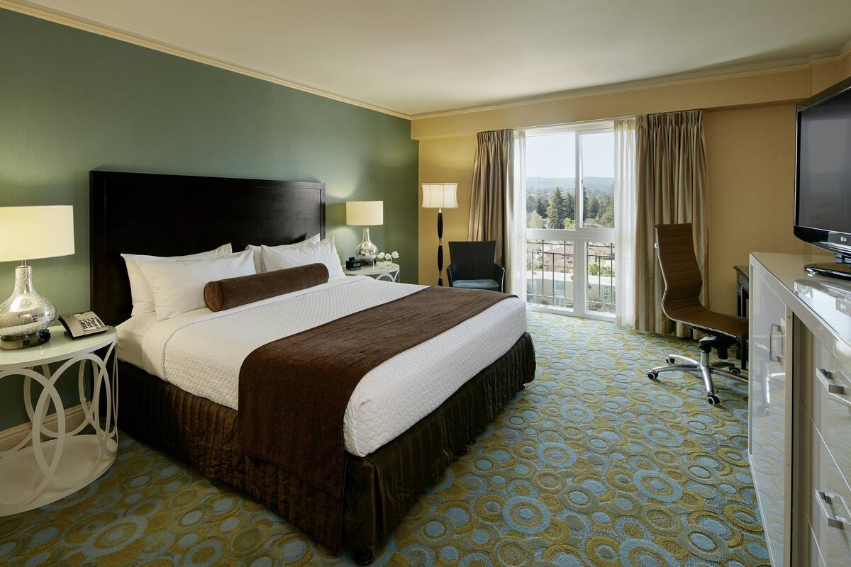 A hotel room with a large bed, two bedside tables with lamps, a desk with a chair, a television, and a view of the outdoors through a large window.