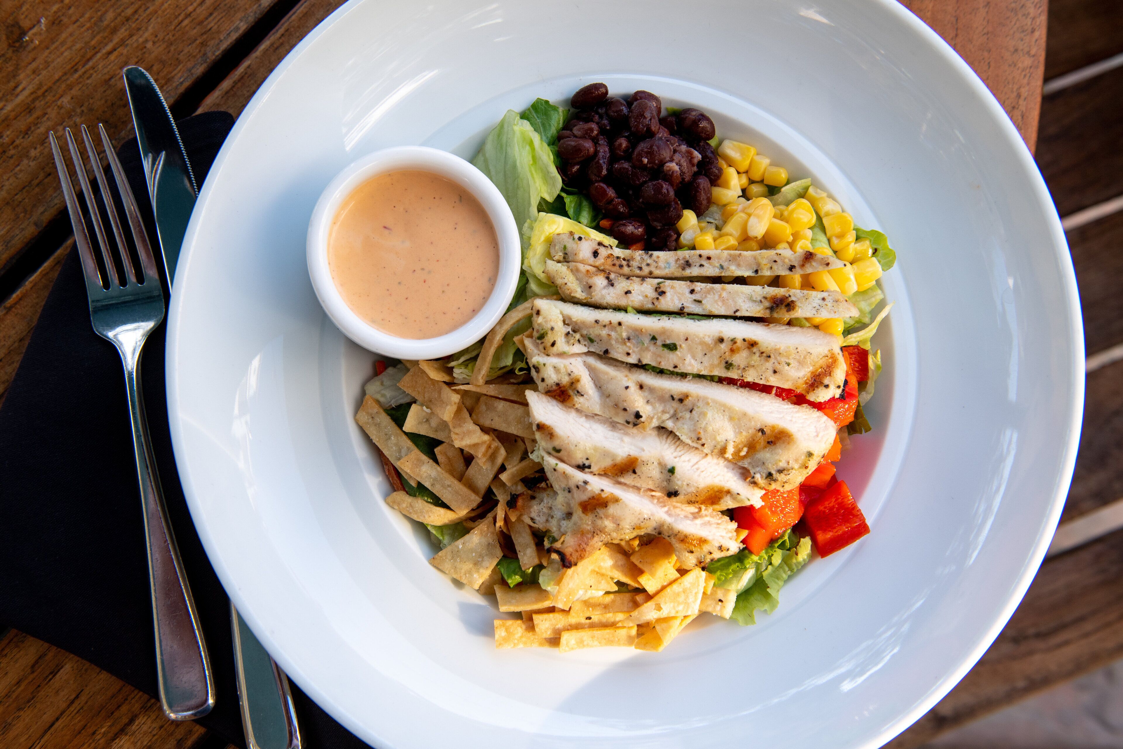 A white plate with a salad containing lettuce, corn, red bell peppers, beans, and tortilla strips. There is a small bowl of dressing on the side.