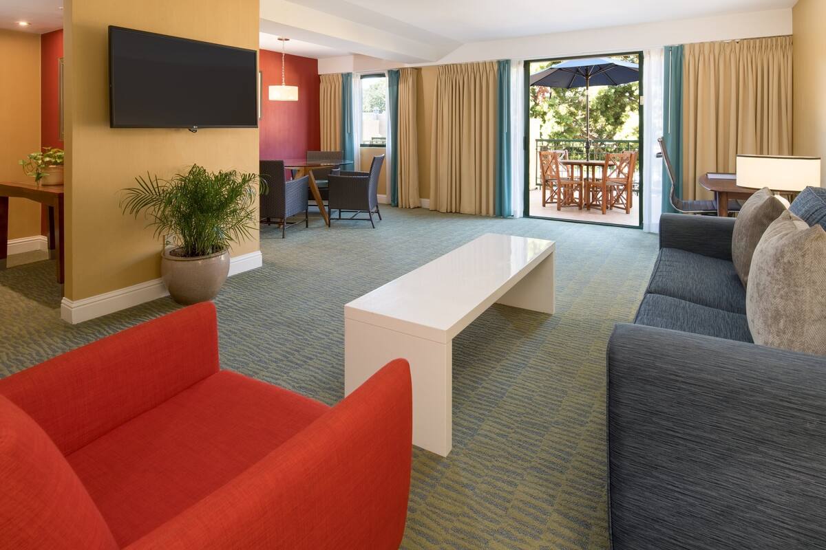 A hotel room with a living area featuring a sofa, a red armchair, a white coffee table, a flat-screen TV mounted on the wall, and a dining area with a table and chairs.