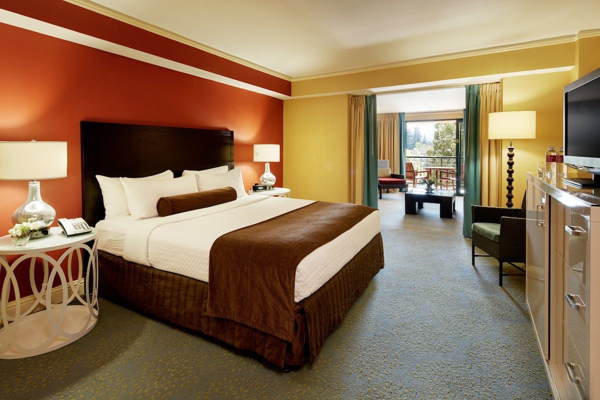 A hotel room with a large bed, two bedside tables with lamps, a TV, and a balcony with seating.