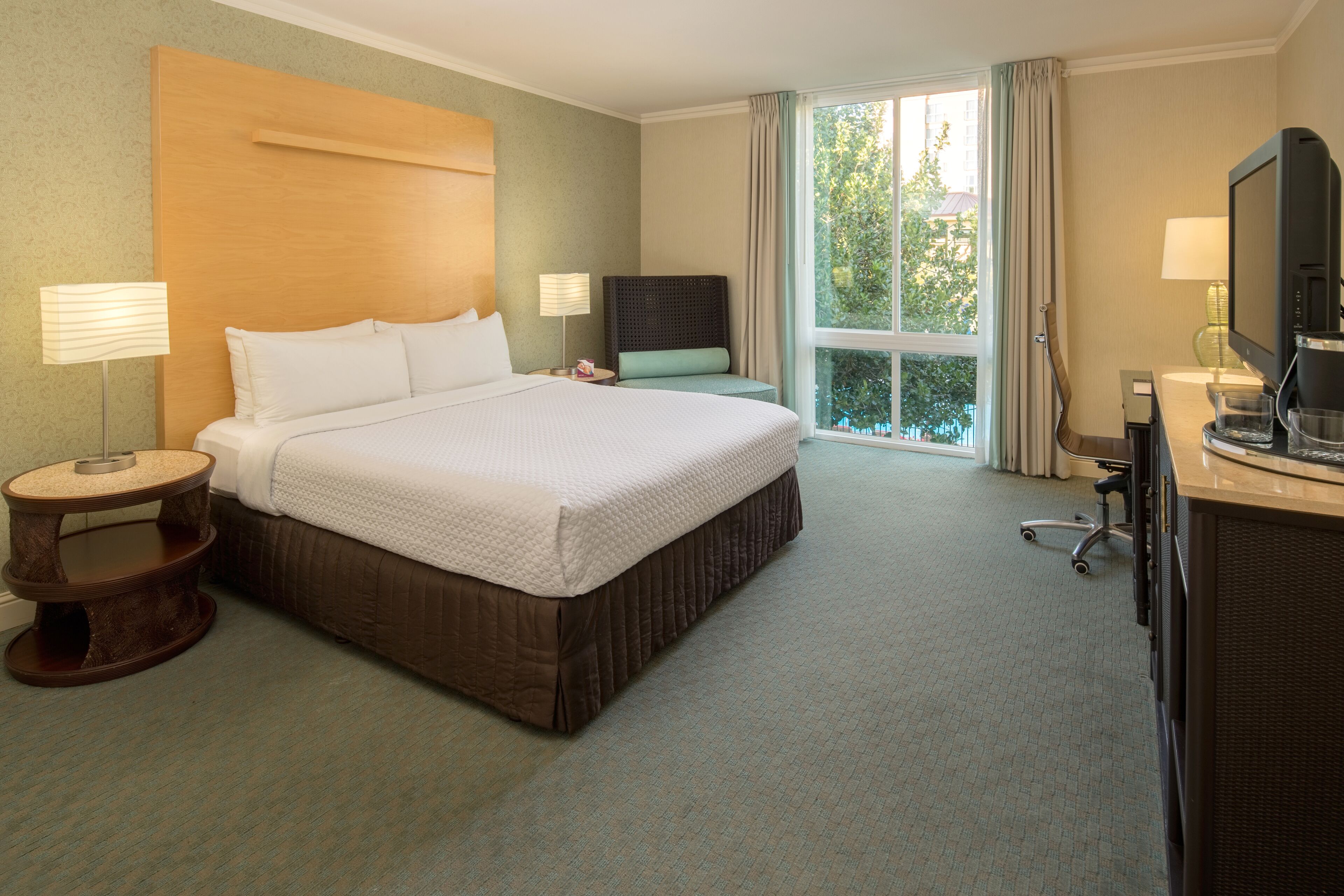 A hotel room with a large bed, a desk with a computer, a chair, a lamp, and a window with curtains.