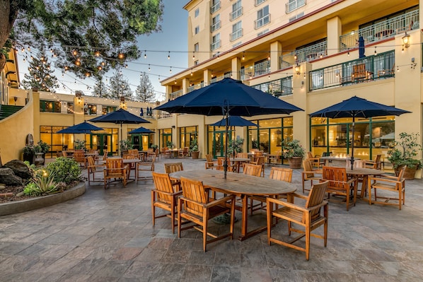 An outdoor patio with wooden tables and chairs, surrounded by potted plants and string lights.