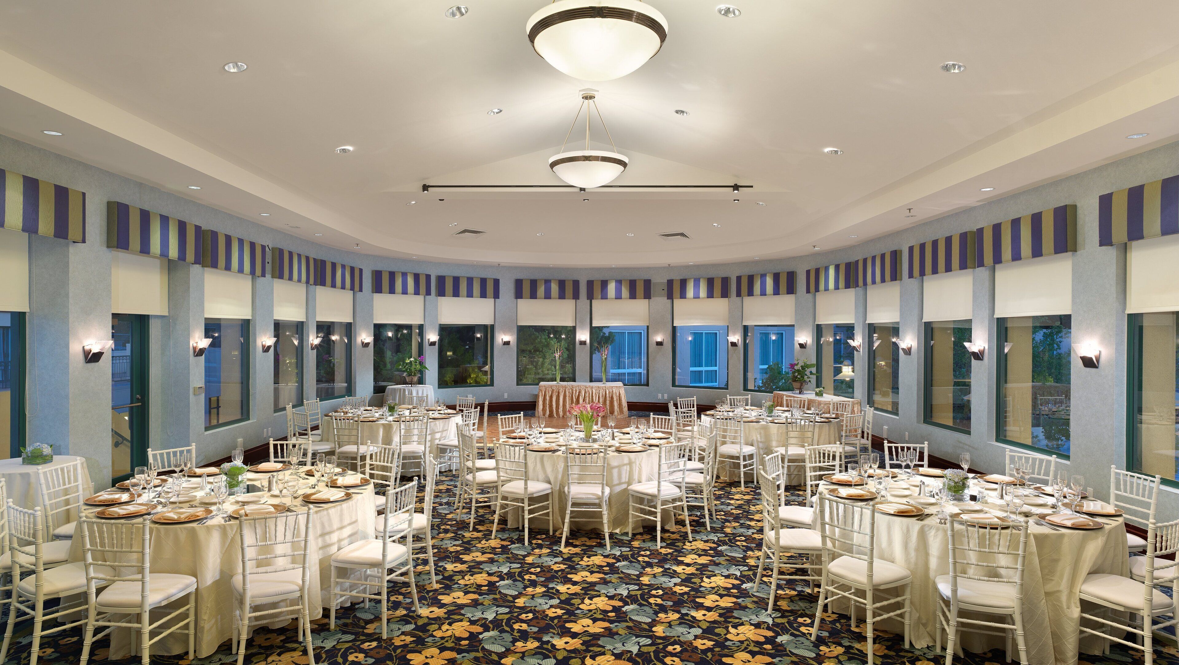 A banquet hall with round tables set for a formal event, featuring a central stage and large windows.