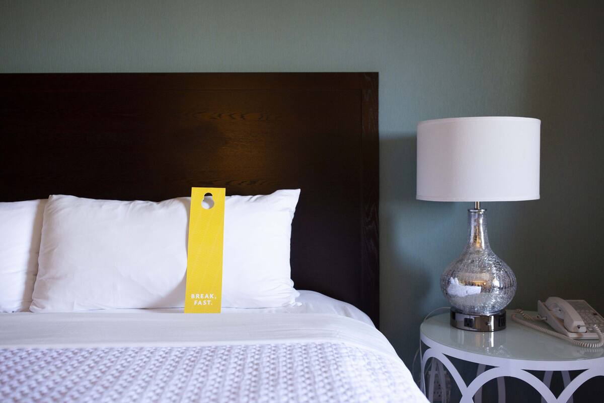 A neatly made bed with white linens, a yellow tag, and a white lamp on a small white table beside it.
