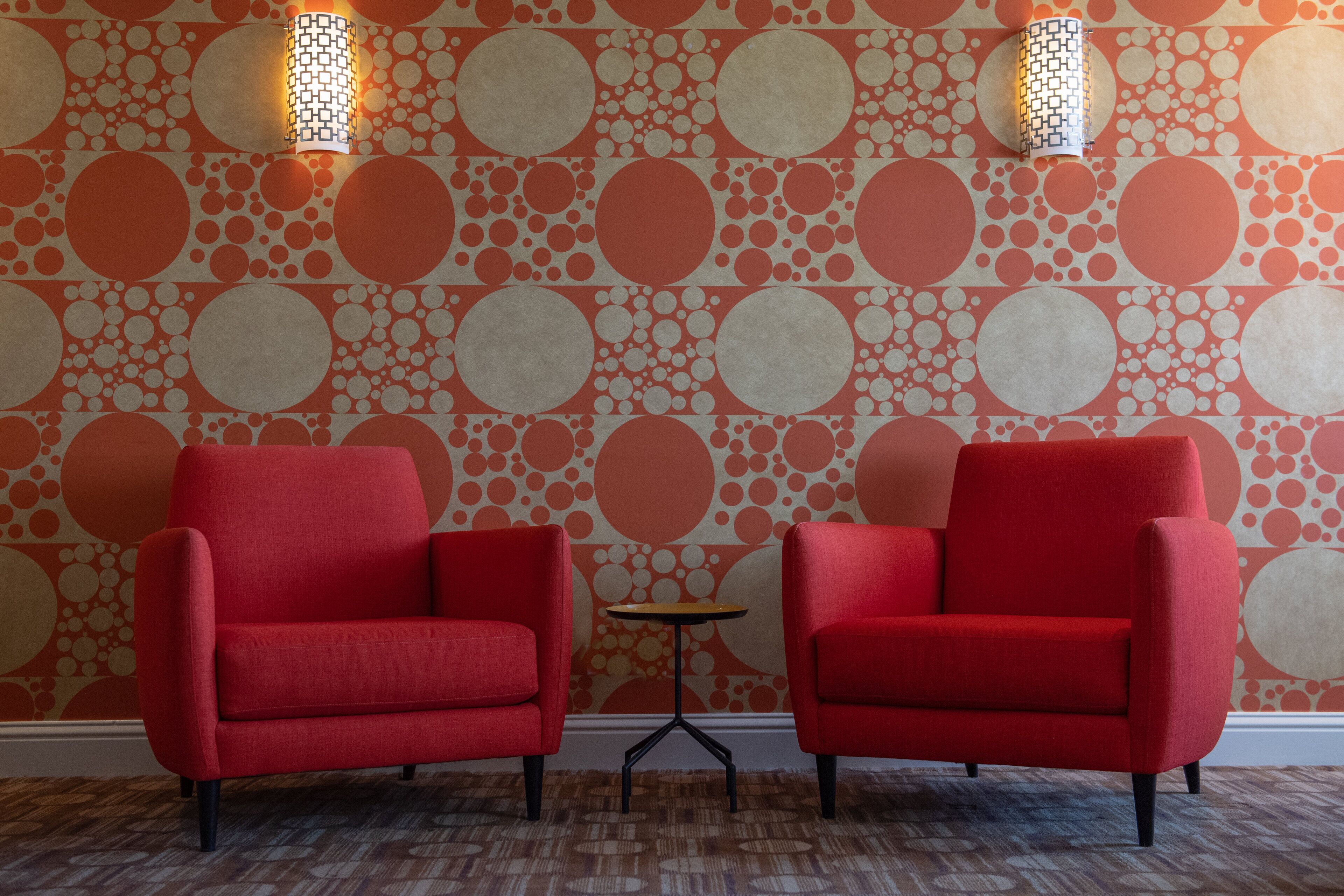 Una habitación con dos sillones rojos y una pequeña mesa, con papel tapiz estampado y dos luces fijadas a la pared.