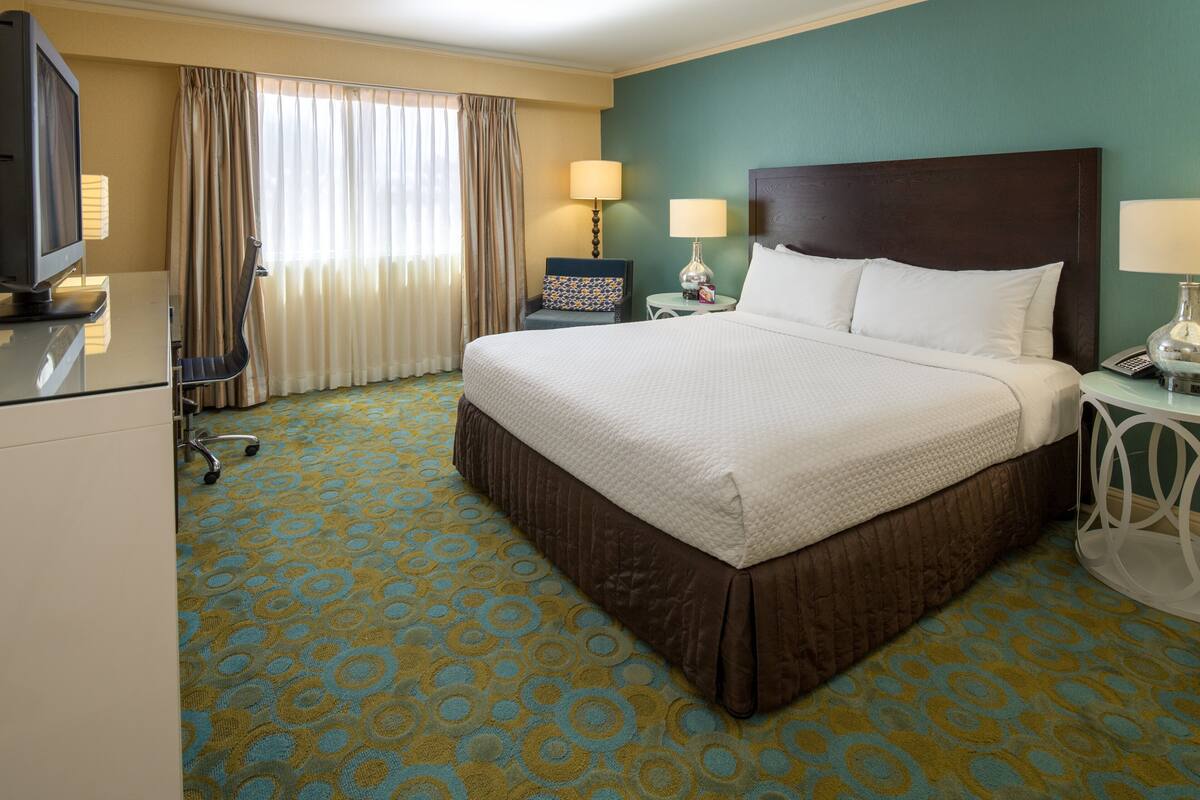 A hotel room with a large bed, a desk with a television, a chair, and a nightstand with a lamp.