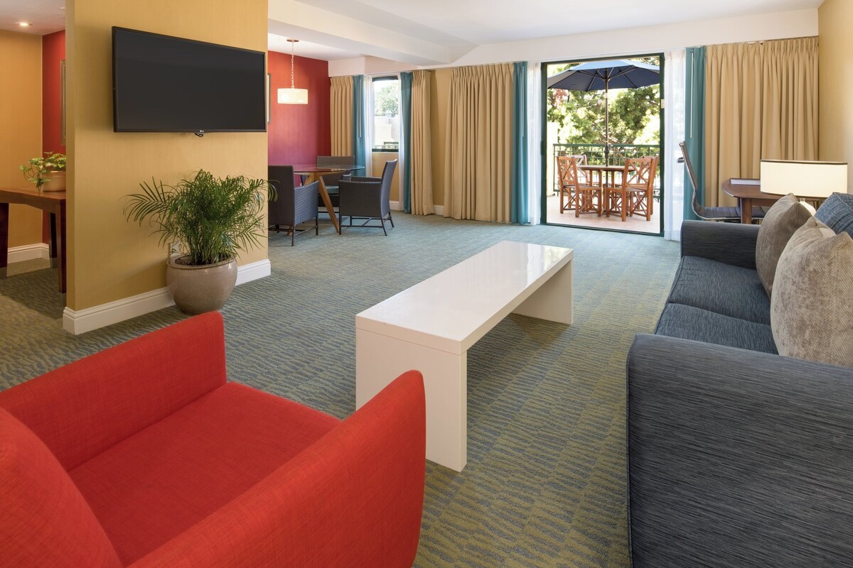 Hotelkamer met een zithoek, bestaande uit een bank, een rode fauteuil, een witte salontafel, een aan de muur bevestigde flatscreen-tv en een eethoek met een tafel en stoelen.