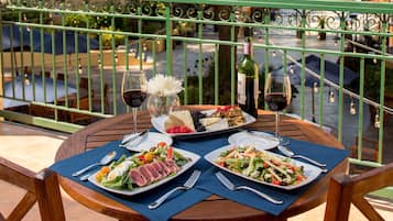A round wooden table set with plates of food, wine glasses, and a bottle of wine on a balcony with a green railing.