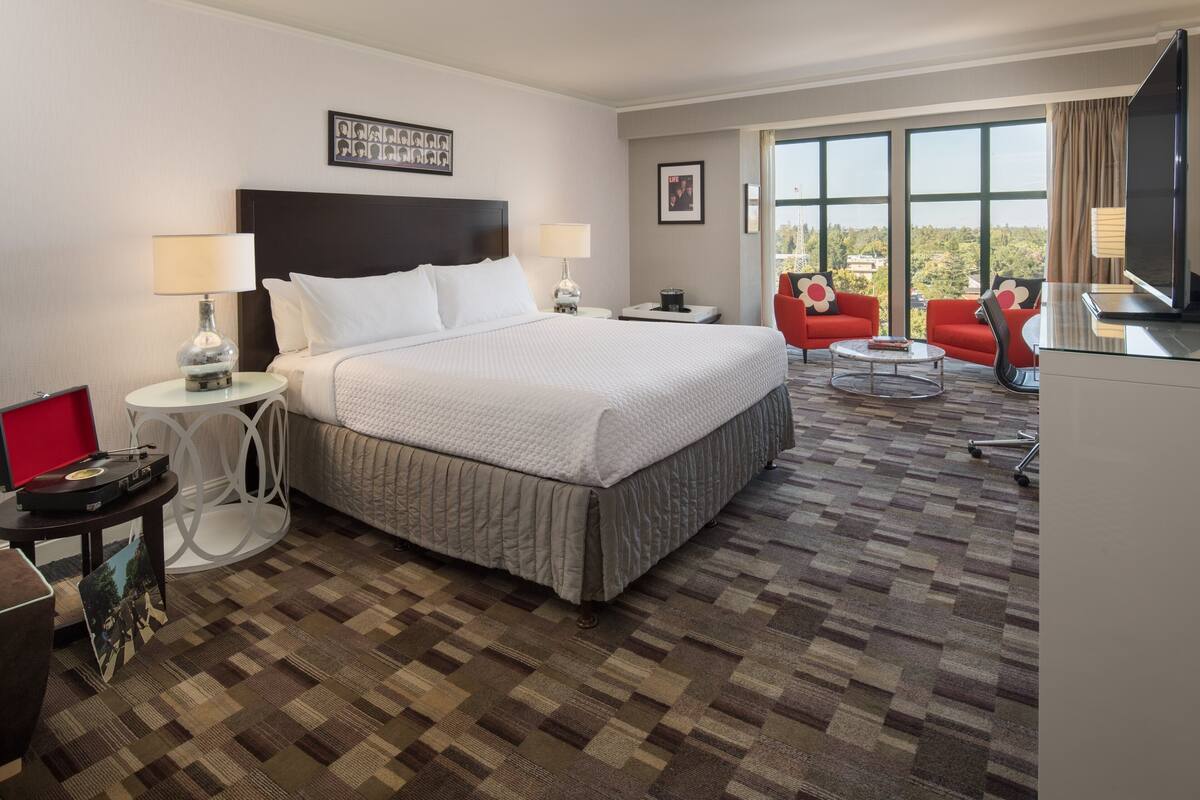 A hotel room with a large bed, two bedside tables, a TV, a red armchair, and a view of the outdoors.