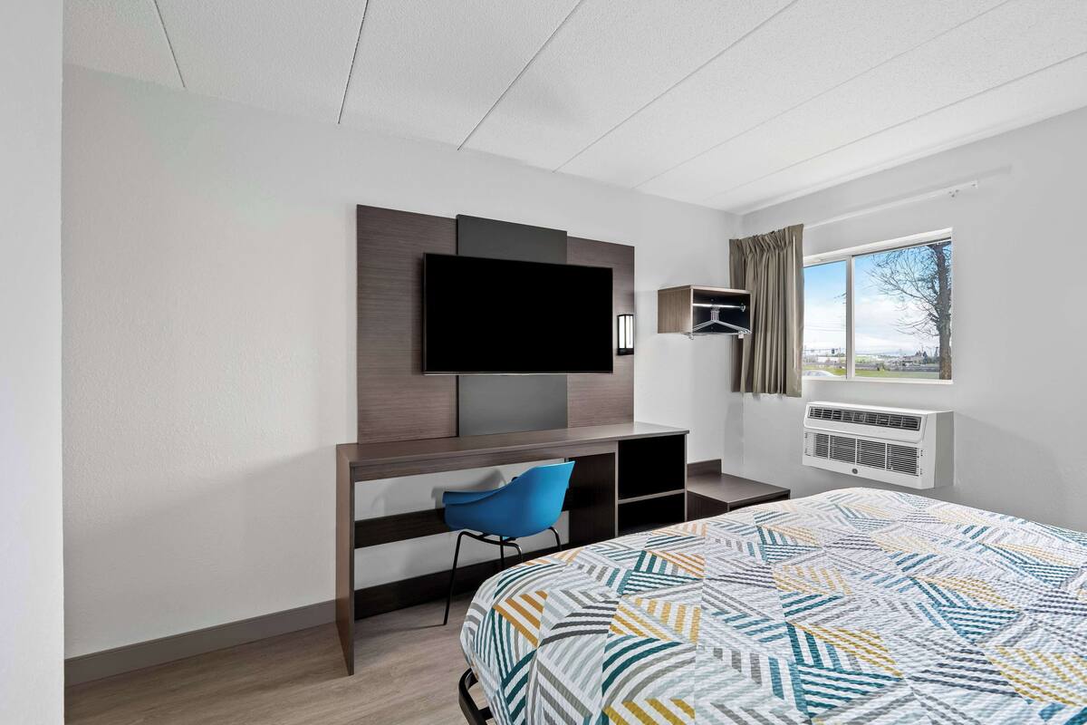 A modern hotel room with a large bed, a desk with a chair, a flat-screen TV mounted on the wall, a window with a view, and an air conditioning unit.