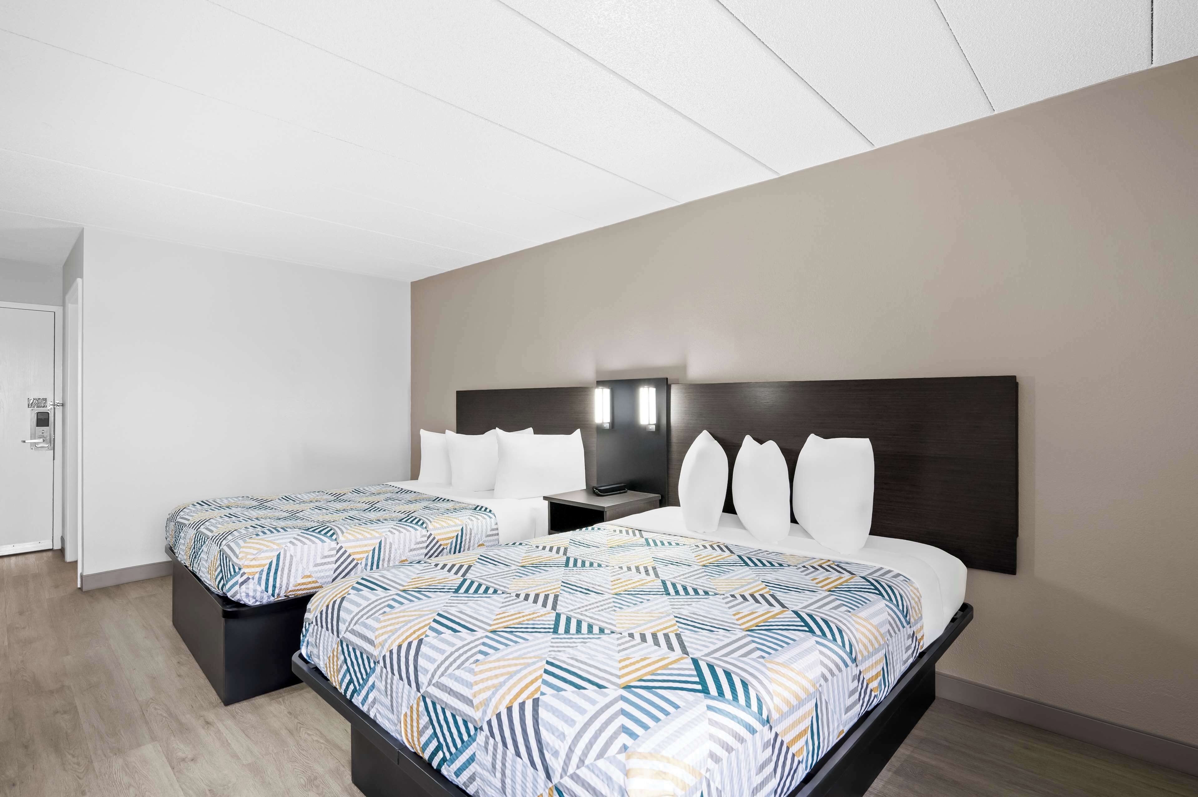 A hotel room with two beds, a dark headboard, a nightstand, and a wall-mounted light fixture.