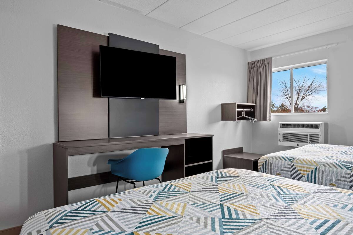 A hotel room with two beds, a flat-screen TV, a desk with a chair, a wardrobe, and a window with a view of trees.