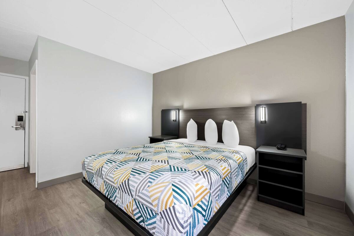 A modern hotel room with a large bed, a headboard with integrated lighting, a bedside table, and a built-in shelf unit.