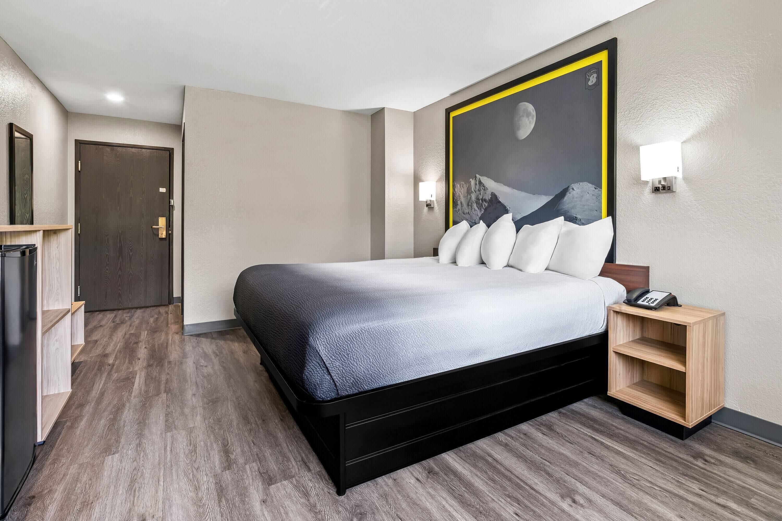 Une chambre d’hôtel avec un lit, une table de chevet, une lampe fixée au mur et un tableau représentant un paysage de montagne.