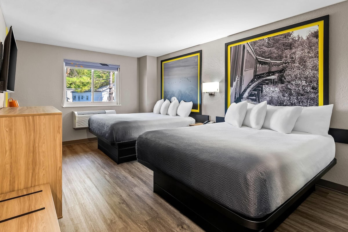 Ein Hotelzimmer mit zwei Betten, einem Holztisch, einem Fenster mit Blick auf Häuser und einem großen gerahmten Bild an der Wand.