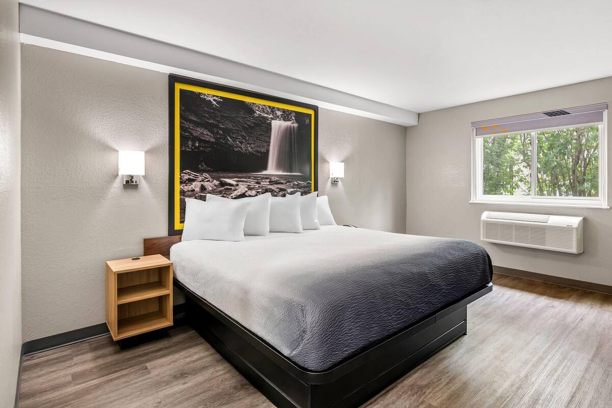 Ein modernes Hotelzimmer mit einem großen Bett, einem Nachttisch, einer Wand-Klimaanlage und einem Fenster mit Blick auf Bäume.