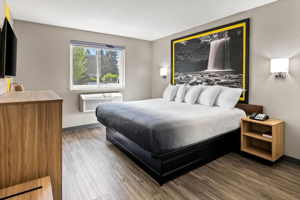 Ein modernes Hotelzimmer mit einem großen Bett, einem Schreibtisch, einem Stuhl, einem Fenster mit Blick auf Bäume und einer Wandleuchte.