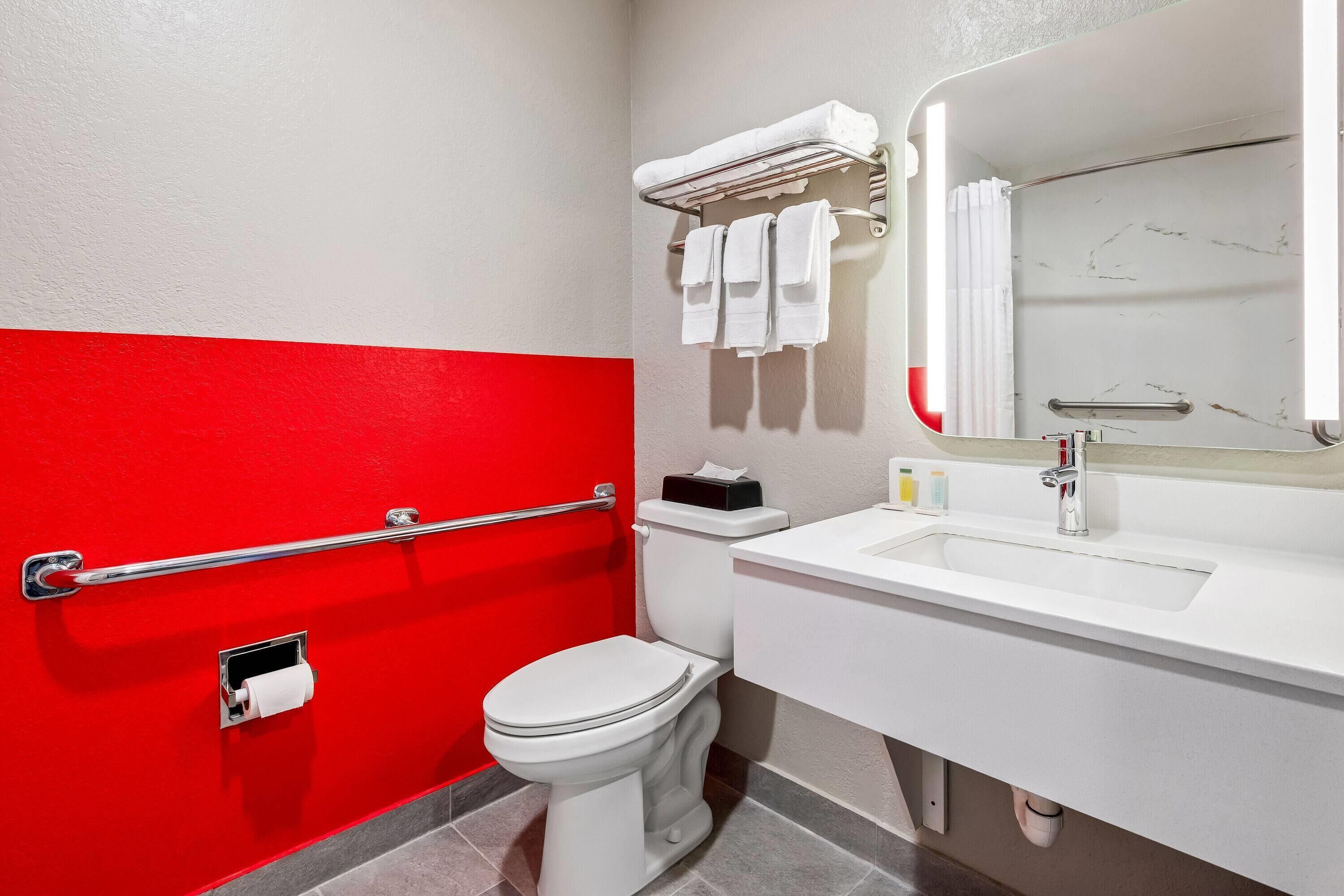 Un baño con pared roja y blanca, un inodoro blanco, un lavabo con espejo y un perchero con toallas.