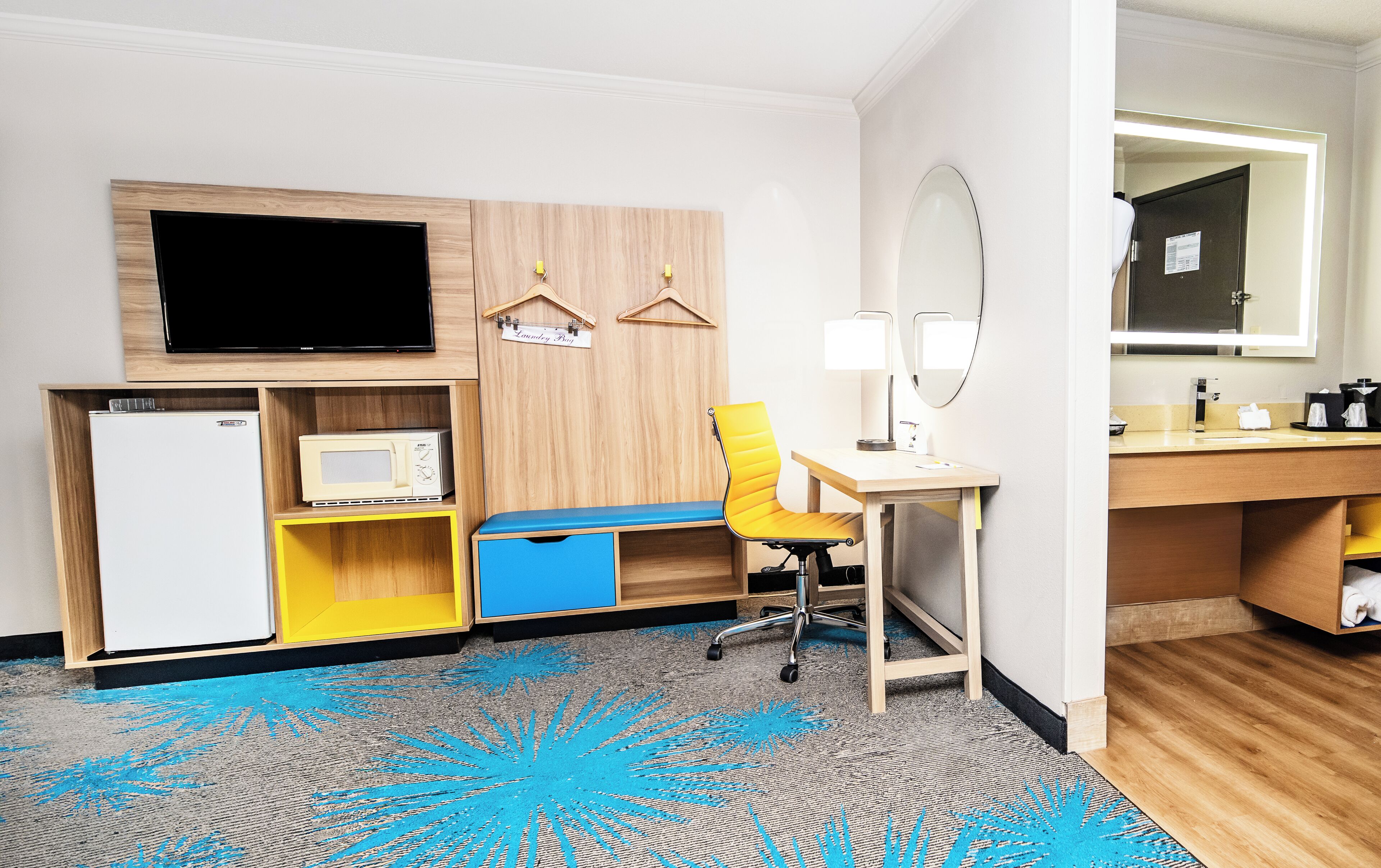 Habitación de hotel con televisión, microondas, escritorio con silla, espejo y una pequeña kitchenette.
