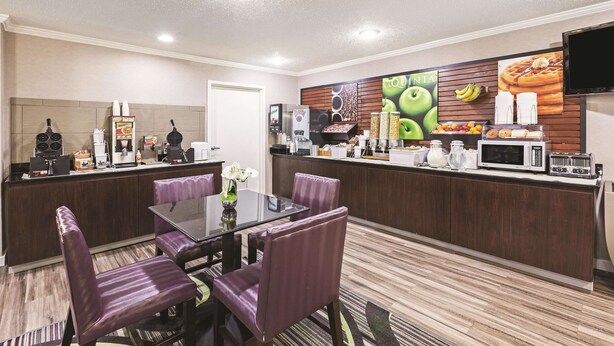 Un comedor de hotel con una mesa de vidrio, sillas tapizadas de color púrpura, un mostrador de buffet con alimentos, una máquina de café y un televisor de pantalla plana montado en la pared.