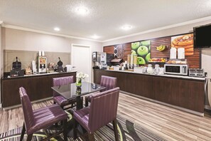 Un comedor de hotel con una mesa de vidrio, sillas tapizadas de color púrpura, un mostrador de buffet con alimentos, una máquina de café y un televisor de pantalla plana montado en la pared.