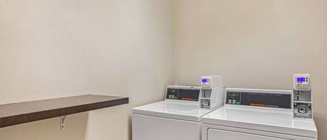A laundry area with two white commercial dryers and a wooden shelf.