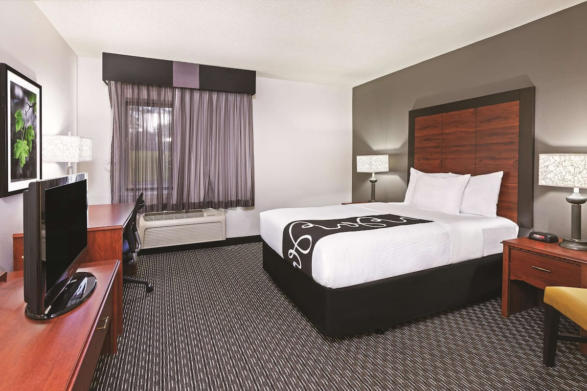 A hotel room with a large bed, a desk with a flat-screen TV, a chair, two bedside lamps, and a window with curtains.