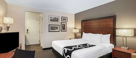 A hotel room with a large bed, a desk with a chair, a TV, and a door leading to another room.