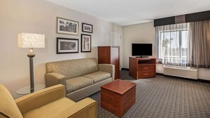 Habitación de hotel con sofá, sillón, una mesita de madera, televisor de pantalla plana, lámpara de pie y un ventanal con cortinas.