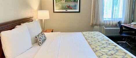 A hotel room with a bed, a desk, a chair, a lamp, and a framed picture of a river scene.