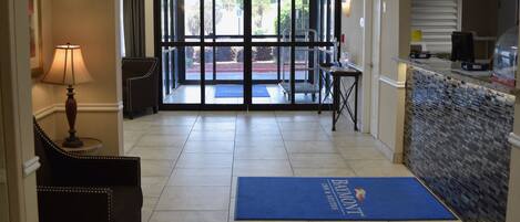 Un vestíbulo de hotel con una mesa de recepción, un felpudo azul con el nombre del hotel, una lámpara y una silla.