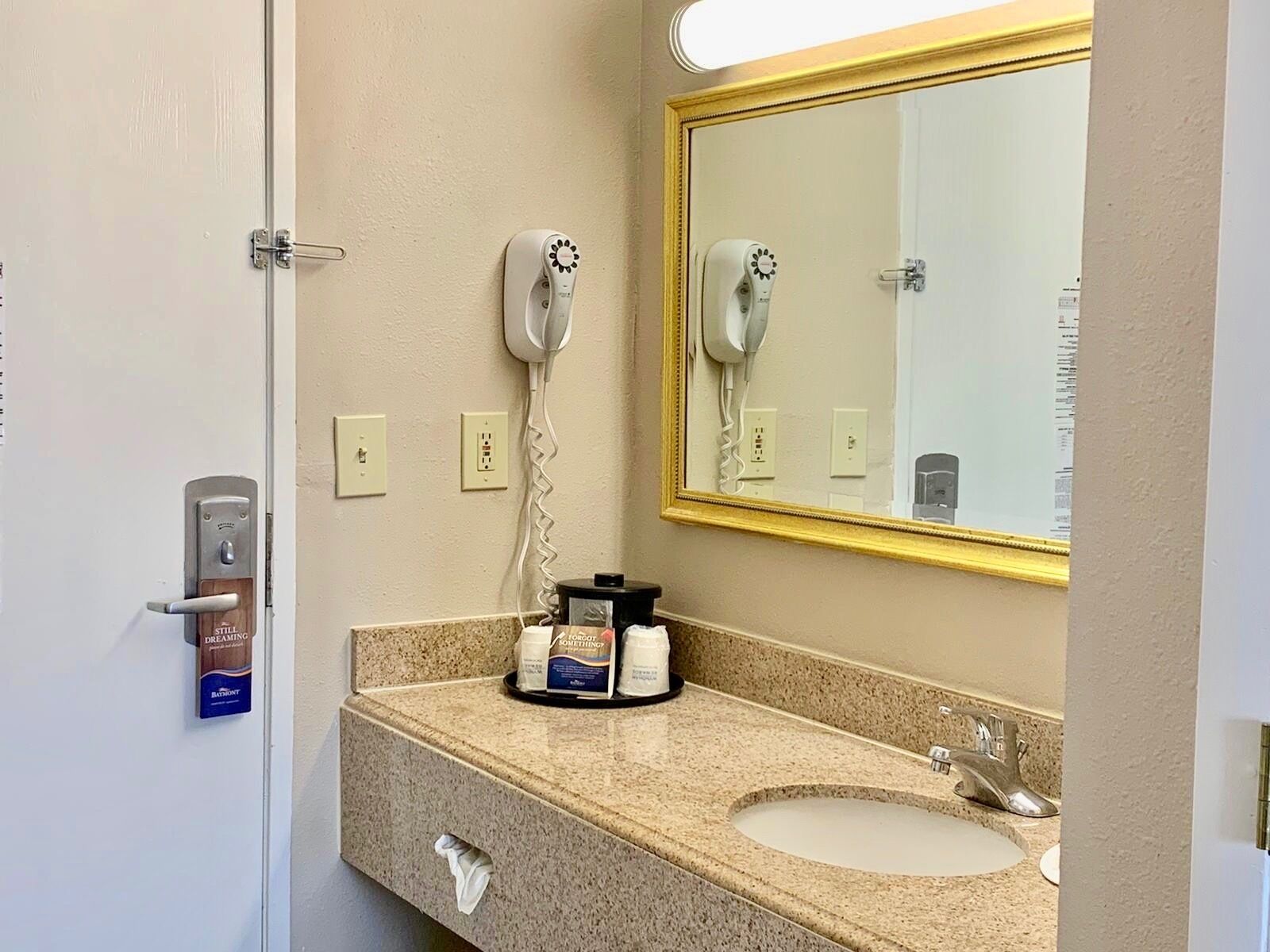 A hotel bathroom with a sink, mirror, hairdryer, and a towel dispenser.