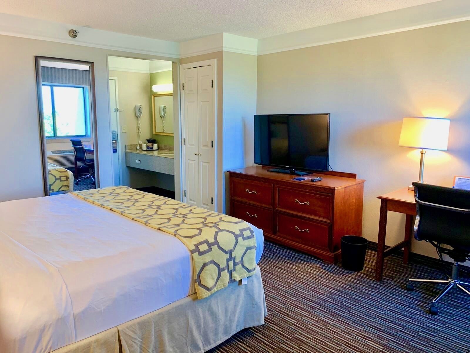 Una habitación de hotel con cama, televisión, escritorio con lámpara, silla y baño visible a través de una puerta abierta.
