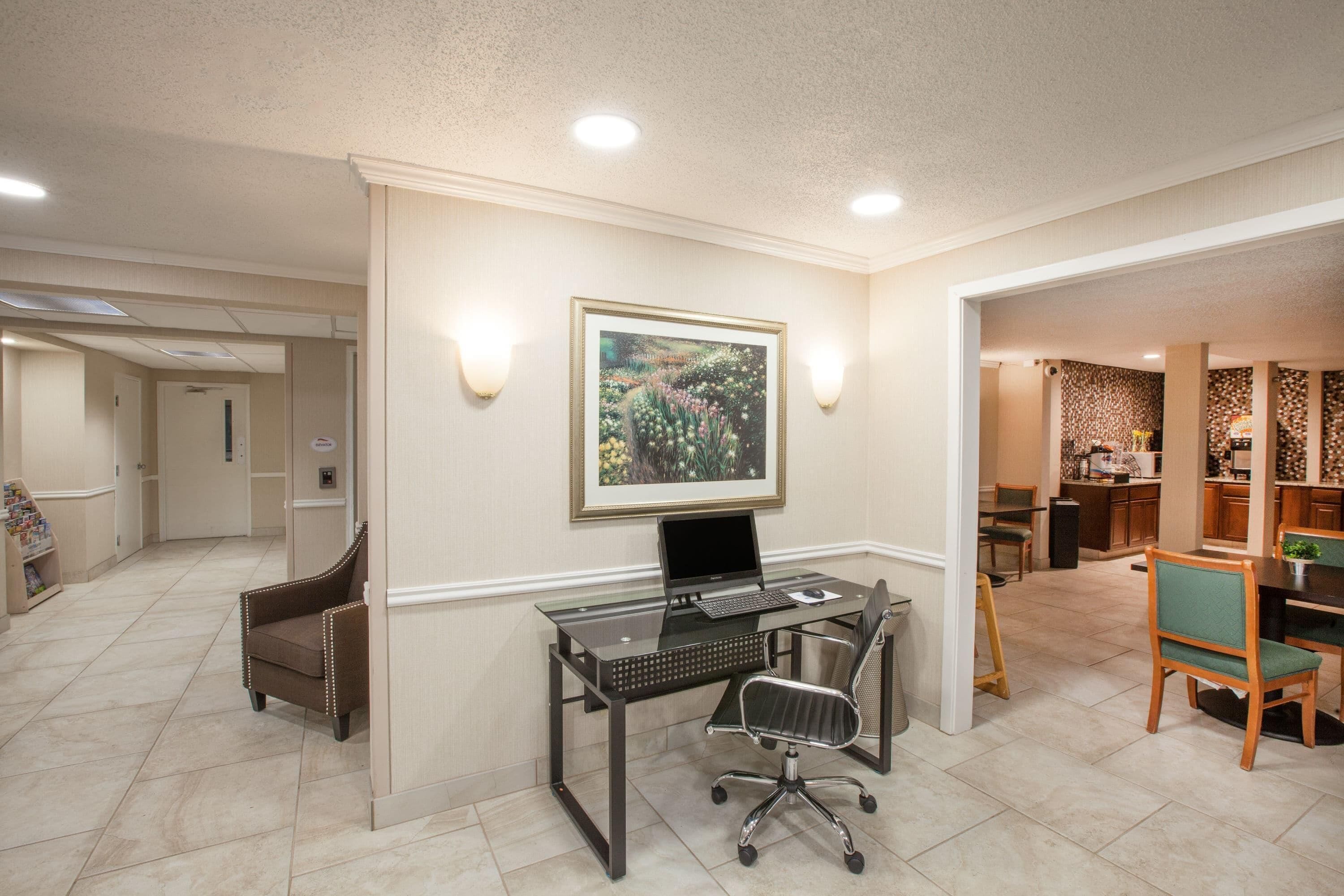 A modern office space with a glass desk, a laptop, a framed picture on the wall, and a dining area with chairs.