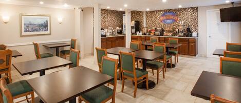 Un comedor con varias mesas y sillas, una zona de cocina con cafetera, y un cuadro en la pared.