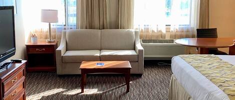 A hotel room with a sofa, a small table, a bed, a TV, a lamp, and a window with curtains.