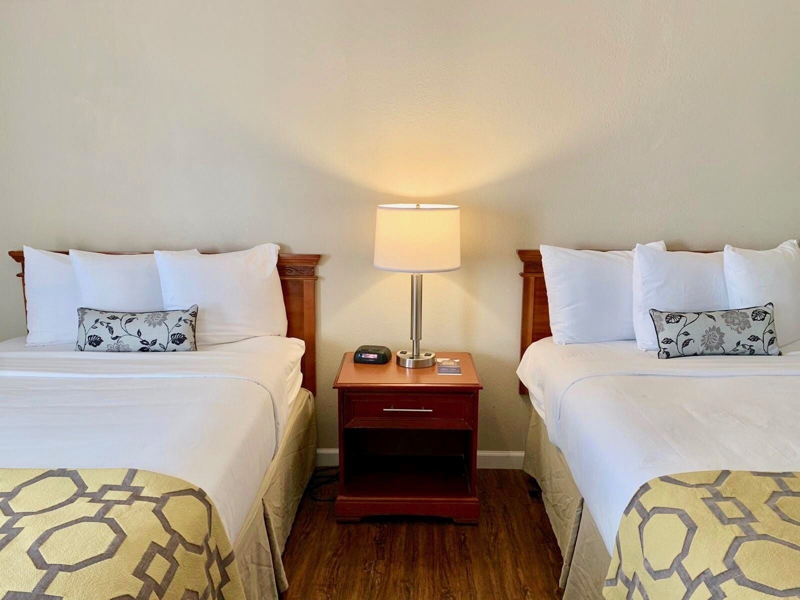 A hotel room with two beds, each with a patterned bedspread, a bedside table with a lamp, and a small electronic device on the table.
