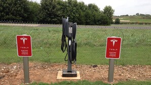 A Tesla electric vehicle charging station with two signs, a grassy area, and a fence in the background. - Super 8 by Wyndham Russellville (Russellville)