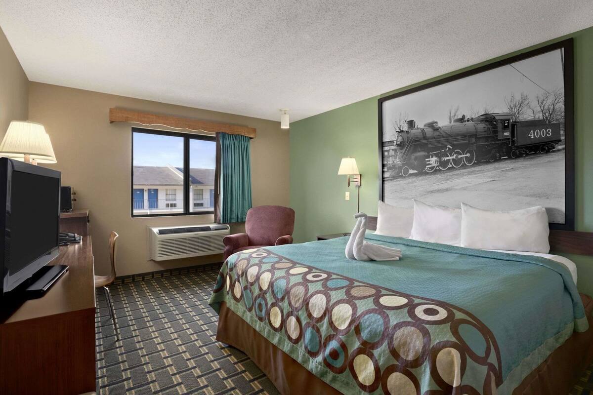 A hotel room with a bed, a TV, a chair, a window with curtains, and a wall-mounted lamp.