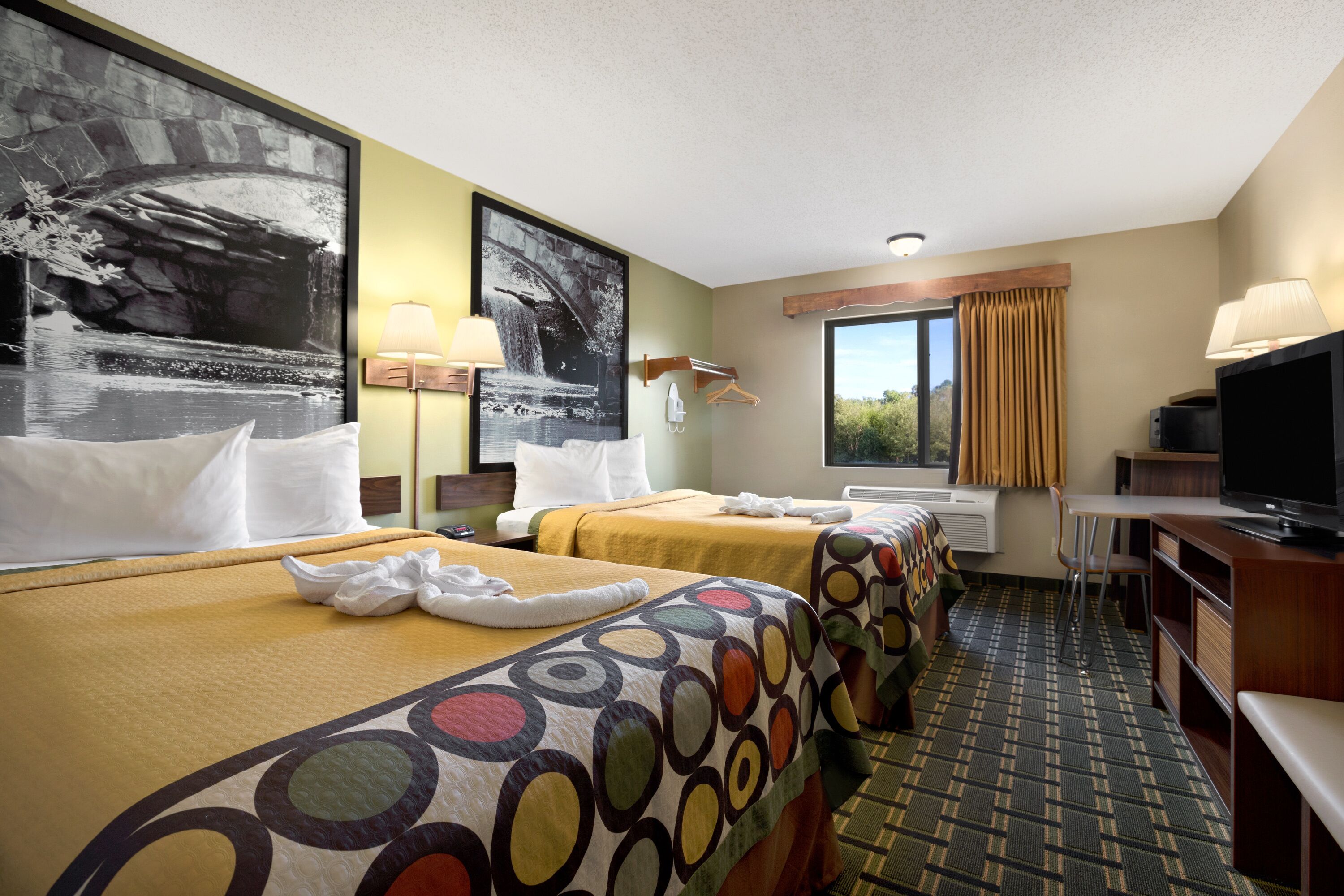 A hotel room with two beds, a TV, a desk, and a window with curtains.