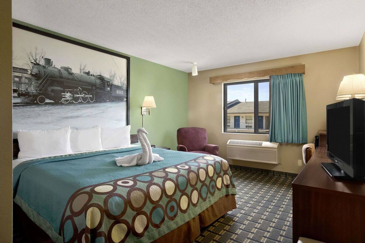 A hotel room with a bed, a chair, a television, a window with curtains, and a wall-mounted lamp.