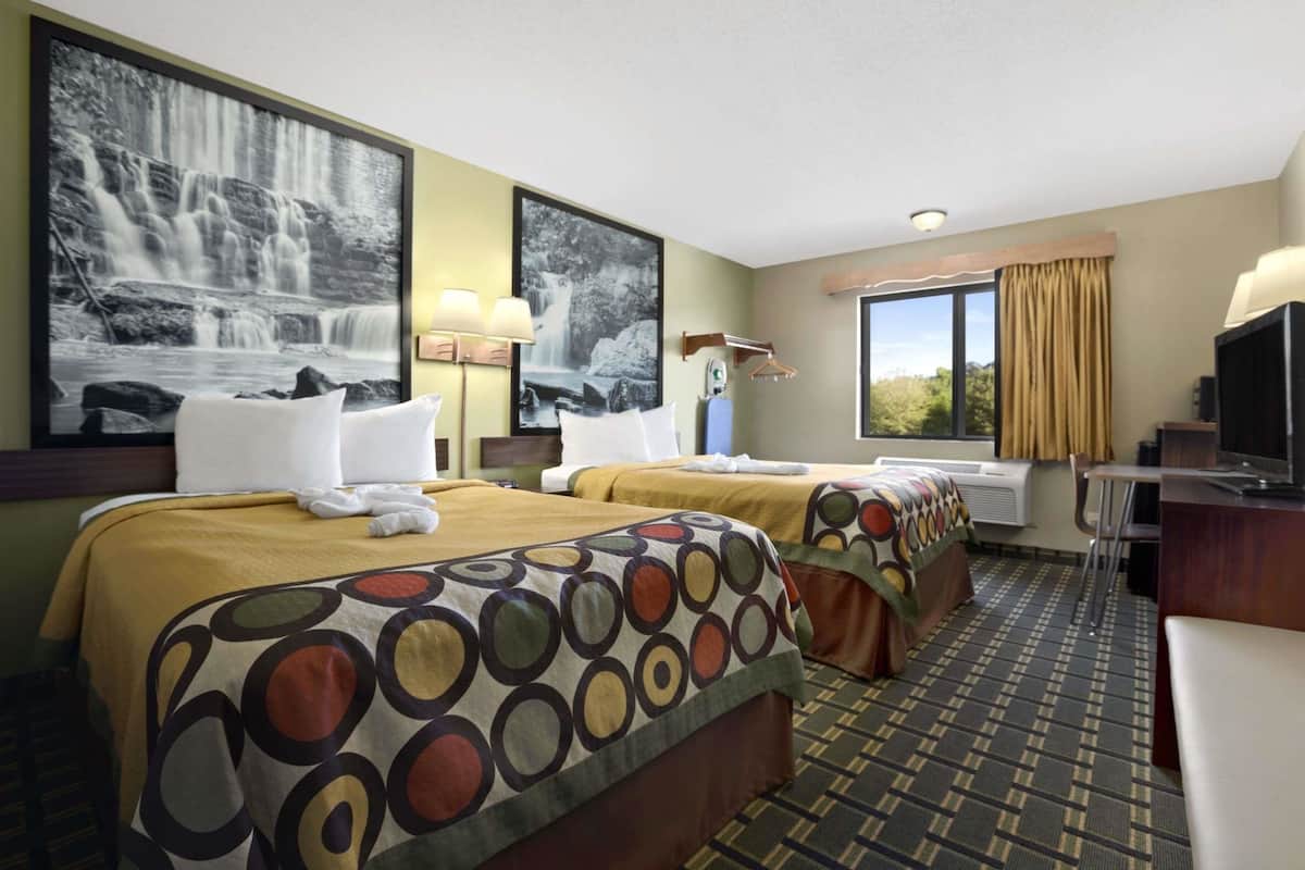 A hotel room with two beds, a desk, a TV, and large windows with curtains.